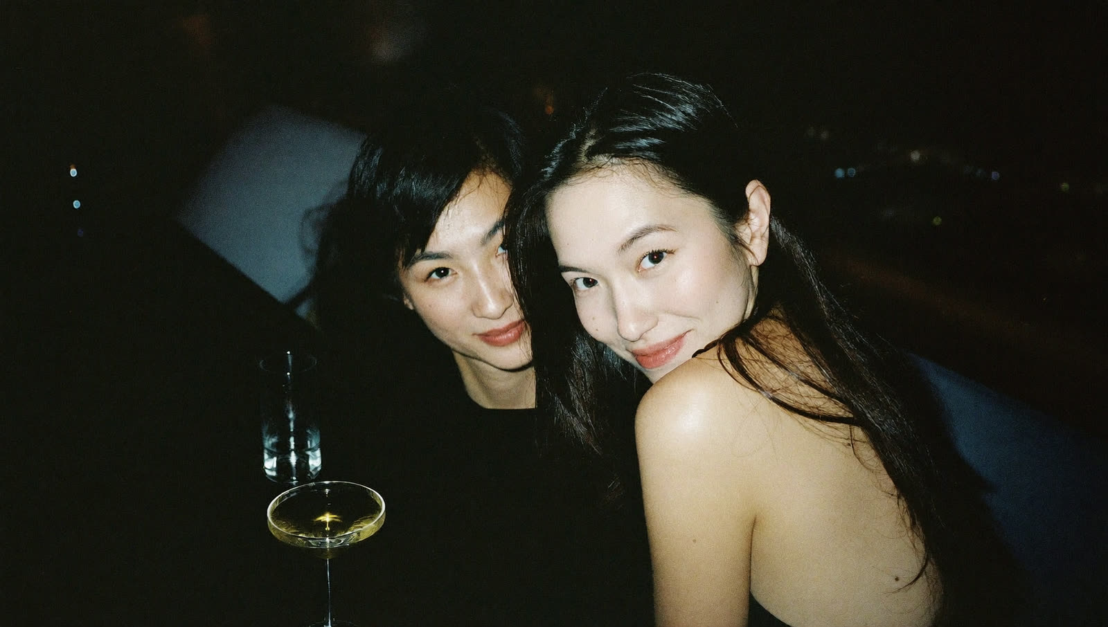 兩位女生在夜晚的店裡靠在一起，桌上放著一杯調酒。